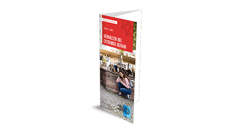 Produktbild Faltblatt Verhalten bei extremer Gefahr Aufrecht stehender Faltflyer mit weißer Überschrift in einem roten Kasten "Verhalten bei extremer Gefahr". Auf der Vorderseite ist eine Person zu sehen, die sich hinter einer Mauer in Deckung befindet.