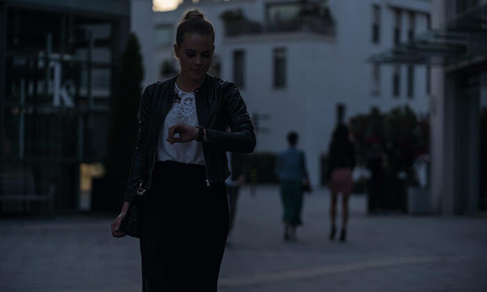 Eine junge Frau steht in der Abenddämmerung in der Innenstadt und schaut auf ihre Armbanduhr.