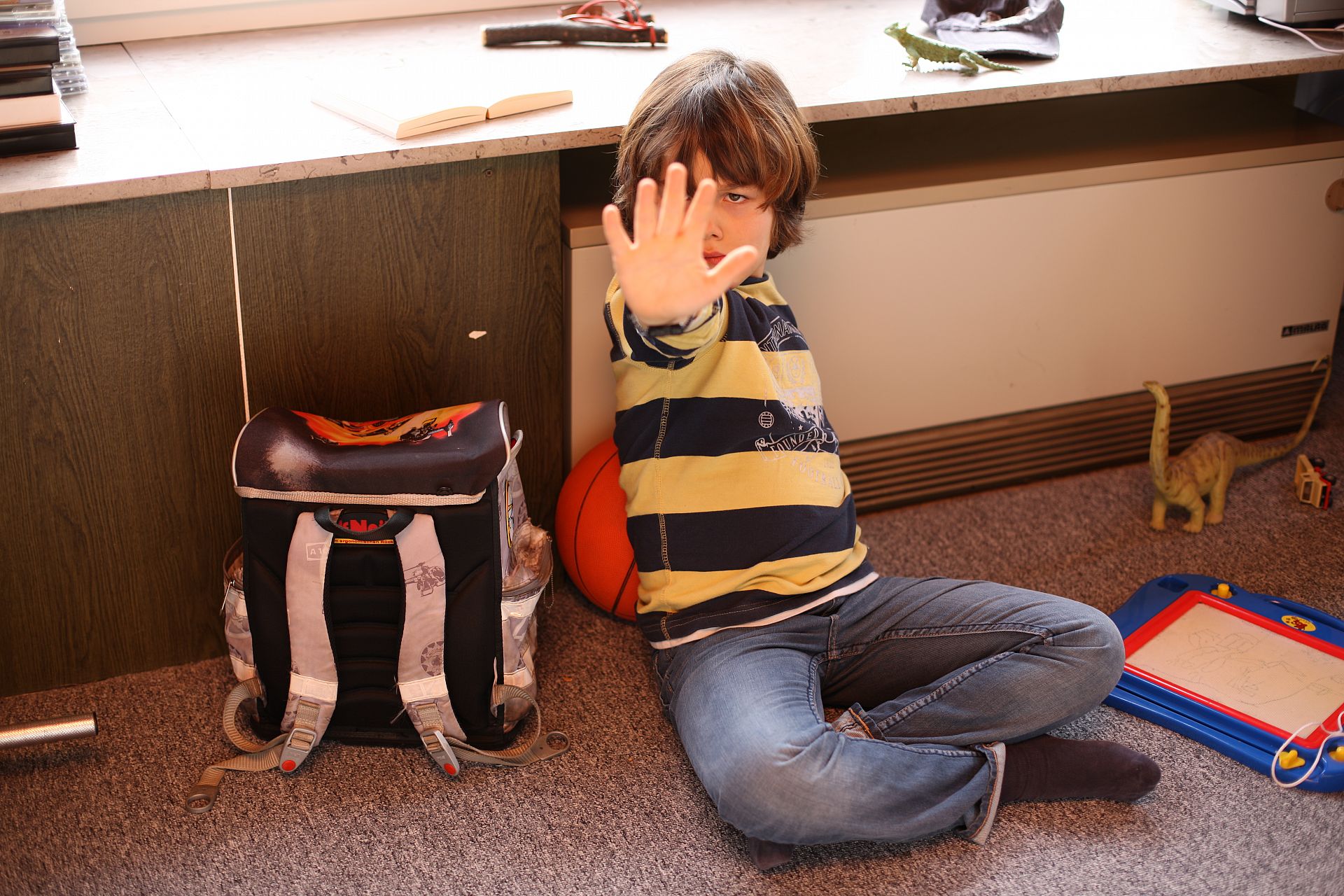 Junge sitzt auf dem Boden neben seinem Schulranzen und zeigt die Stopp-Geste.