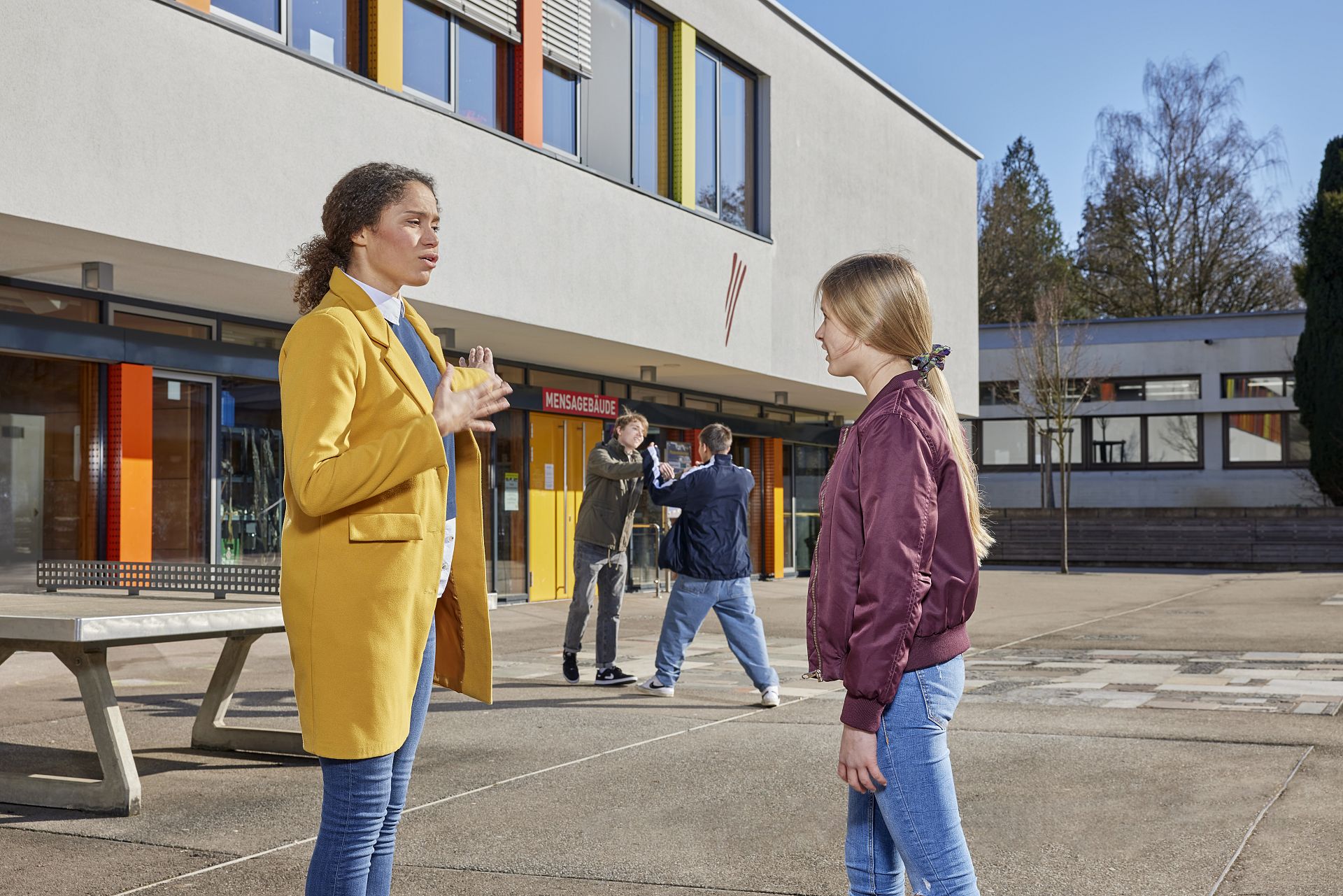 Jungs kämpfen auf dem Schulhof und zwei Mädchen beobachten die Situation.