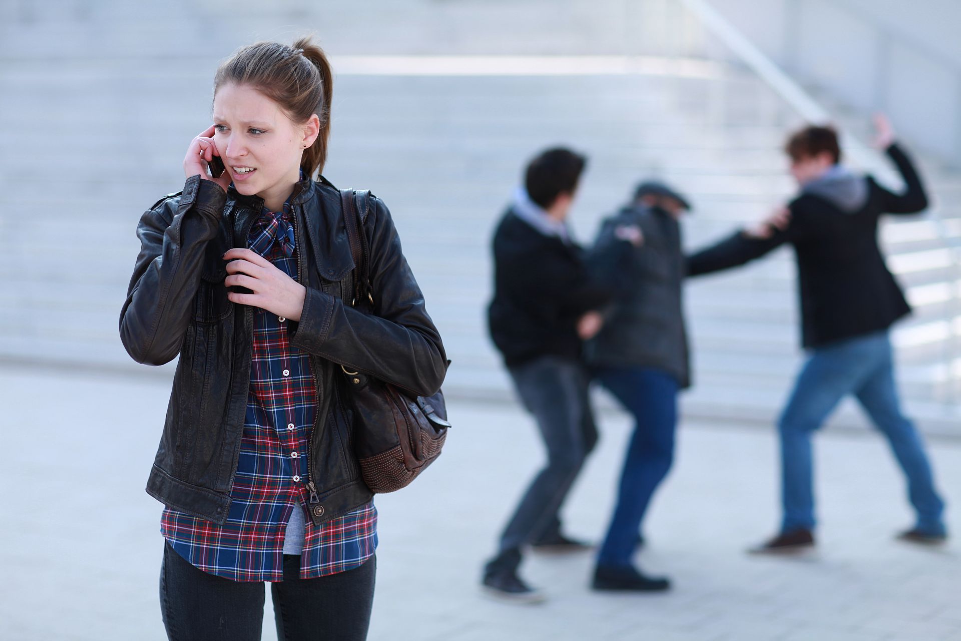 Junge Frau ruft per Smartphone Hilfe. Zwei Männer schlagen auf einen Herr ein.