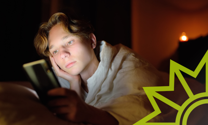 Ein Jugendlicher schaut in einem dunklen Raum, welcher nur mit einem schwachen Licht ausgeleuchtet ist, auf sein Smartphone.