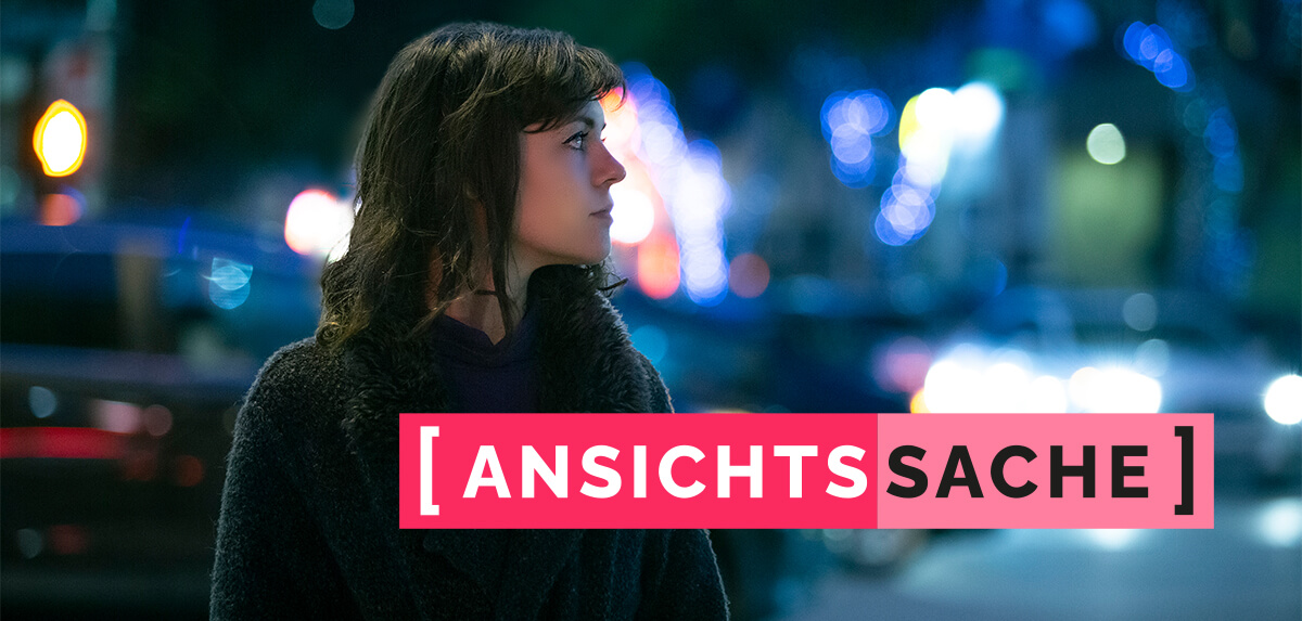 Eine Frau mit dunklen Haaren steht seitlich im nächtlichen Straßenraum und blickt nach links. Im Hintergrund sind unscharfe Lichter von Autos und Straßenbeleuchtung zu sehen. Im Vordergrund der Schriftzug "[Ansichtssache]".