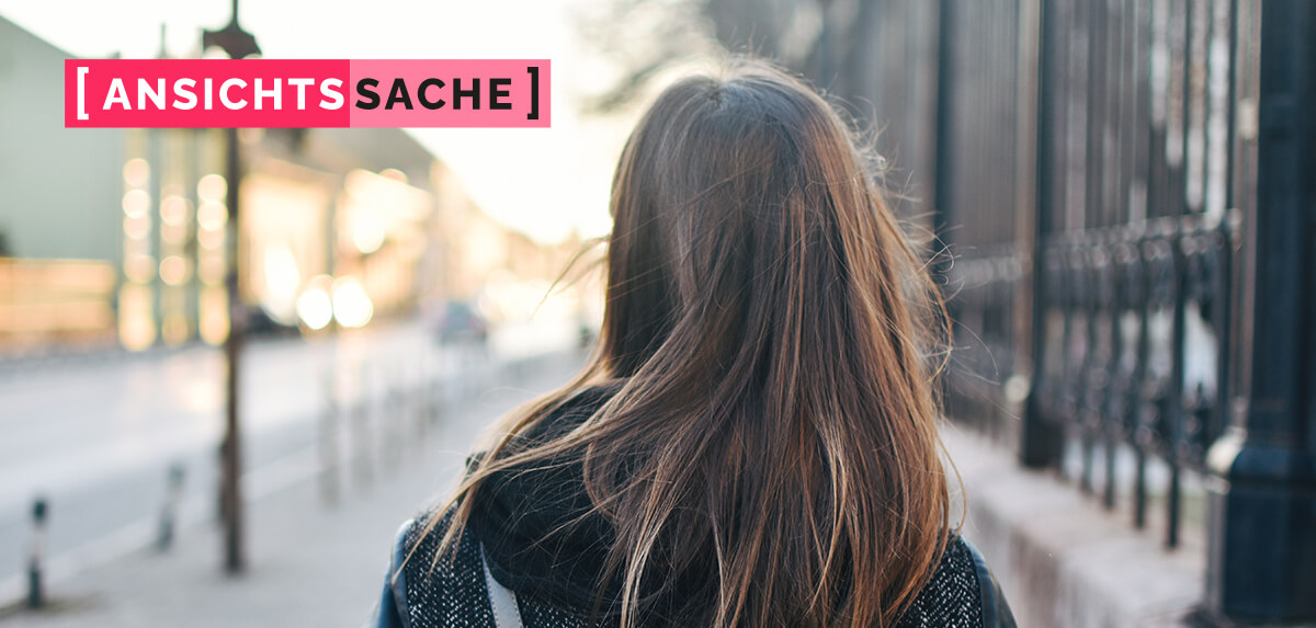 Rückansicht einer Frau mit langen braunen Haaren, die auf einem Gehweg entlang einer Straße und eines Metallzauns geht. Links oben der Schriftzug "[Ansichtssache]".