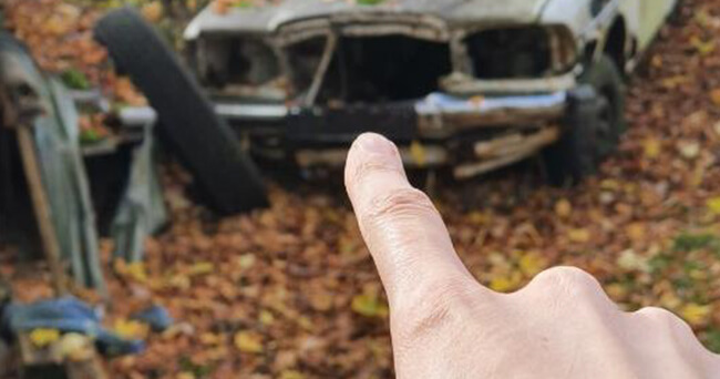 Im Vordergrund zeigt ein ausgestreckter Zeigefinger auf ein stark beschädigtes, verlassenes Auto. Das Fahrzeug steht auf herbstlich bedecktem Boden.
