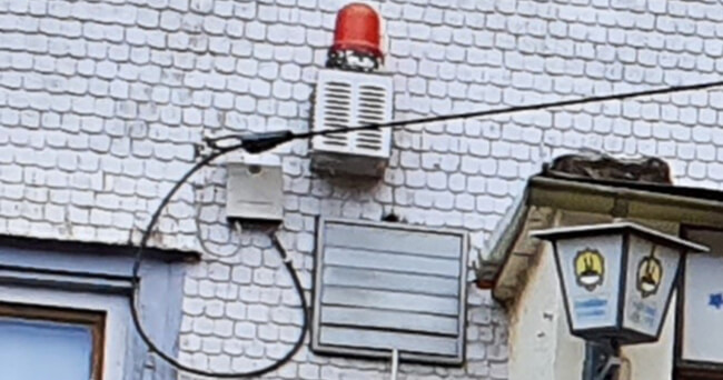 An einer gefliesten Hausfassade ist eine Alarmanlage mit rotem Blinklicht, Lautsprecher und Kabelzuführung befestigt. Darunter sind eine Lampe und weitere technische Elemente sichtbar.