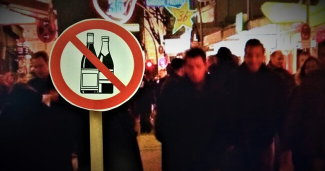 Ein Verbotsschild mit durchgestrichenen Flaschen ist vor einer nächtlich belebten Straße zu sehen, auf der unscharf Menschen vorbeigehen.