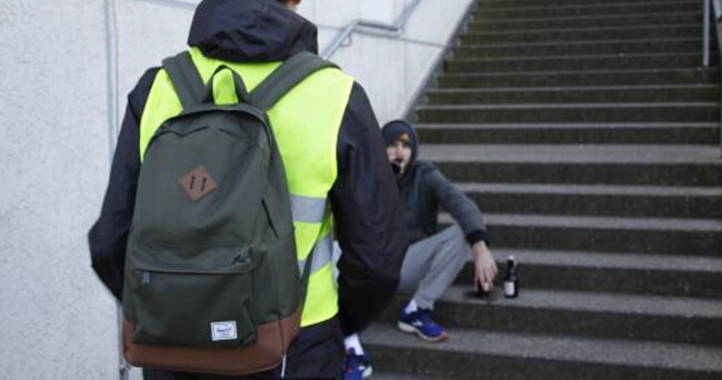 Ein Streetworker mit gelber Warnweste und Rucksack spricht auf einer Außentreppe mit einer jungen Person, die sitzt und eine Bierflasche neben sich stehen hat.