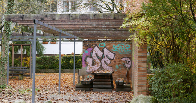 Überdachte Sitzgruppe mit Tisch, umgeben von Bäumen mit Herbstlaub. Eine Backsteinwand im Hintergrund ist mit Graffiti besprüht. Der Platz wirkt ruhig und leicht verwildert, mit Spuren informeller Nutzung.