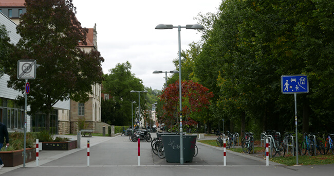 Eine verkehrsberuhigte Straße mit Begrenzungspfosten in der Mitte, Fahrradparkplätzen rechts und Schildern, die eine Spielstraße und das Einfahrtsverbot für Kraftfahrzeuge kennzeichnen. Im Hintergrund sind Menschen zu Fuß unterwegs.