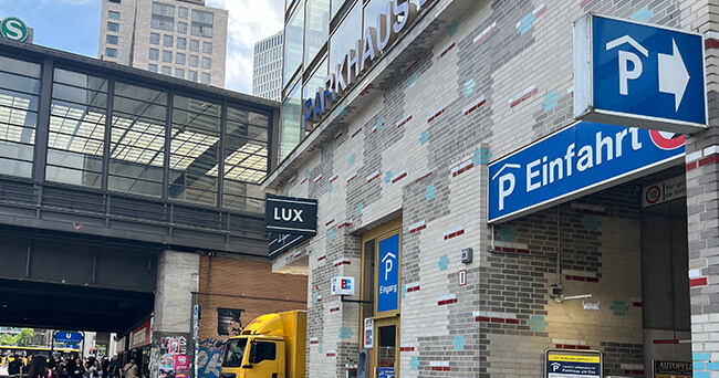 Eingang eines Parkhauses in einer Innenstadt mit mehreren blauen Schildern, die auf Parkmöglichkeiten hinweisen. Links im Bild ist ein S-Bahnhof zu sehen, rechts das Parkhaus mit der Aufschrift „Einfahrt“. Ein gelber Lieferwagen fährt gerade vorbei.