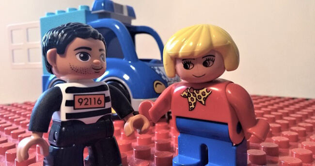 Zwei Legofiguren stehen sich gegenüber, eine mit Sträflingskleidung und eine in Alltagskleidung, im Hintergrund ein Polizeiauto aus Bausteinen.