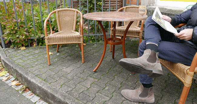 Eine Sitzgruppe aus Holzstühlen und einem Tisch steht auf einem gepflasterten Bereich am Straßenrand. Eine Person sitzt und liest eine Zeitung. Die Umgebung ist ruhig und begrünt.