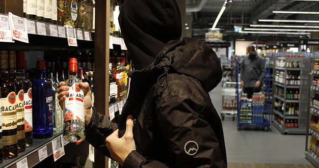 Eine vermummte Person greift in einem Supermarktregal nach einer Flasche Alkohol, während im Hintergrund eine weitere Person zu sehen ist.