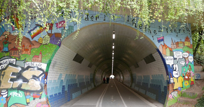 Eine aufgehellte Fußgängerunterführung mit großflächigen Wandgestaltungen und moderner Beleuchtung. Die Wände sind mit bunten geometrischen Formen bemalt, der Boden ist sauber, und die Decke wird durch lineare Lichtquellen erhellt.