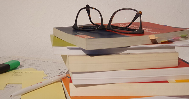 Ein Stapel Fachbücher und Unterlagen mit farbigen Markierungen liegt auf dem Tisch. Obenauf liegt eine Brille. Im Hintergrund liegen Notizzettel und ein Textmarker und ein Kugelschreiber.
