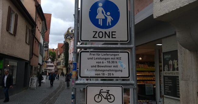 Verkehrsschilder in einer verkehrsberuhigten Innenstadtzone mit Hinweis auf Lieferverkehrszeiten, Fahrrad- und Fußgängerbereich. Im Hintergrund eine belebte Einkaufsstraße mit Menschen.