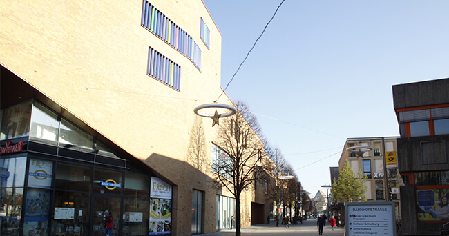 Städtischer Straßenbereich mit moderner Architektur, Sitzgelegenheiten und Fußgängerzone, der durch klare Linien und helle Gestaltung zur Aufenthaltsqualität beiträgt.