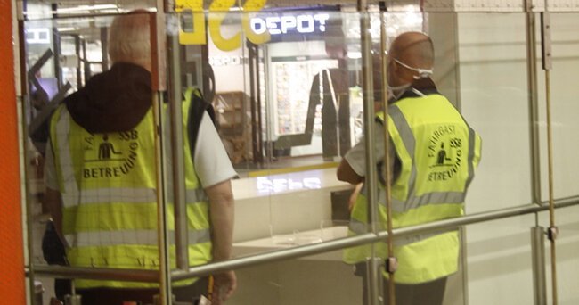 Zwei Personen in gelben Warnwesten mit der Aufschrift „Betreuung“ stehen in einem Bahnhofsumfeld und beobachten den Bereich vor einem Geschäft.