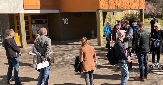 Mehrere Personen stehen vor einem Mehrfamilienhaus mit oranger Fassade und der Hausnummer 10. Einige tragen Jacken oder haben Unterlagen dabei. Im Hintergrund sind weitere Gebäude und Bäume zu sehen. Die Szene wirkt wie eine geführte Ortsbegehung an einem sonnigen Tag.