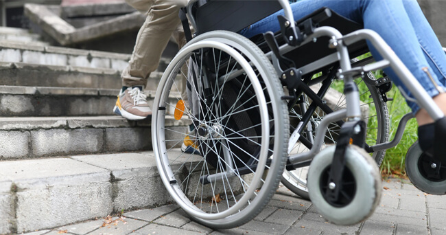 Ein Rollstuhlfahrer wird von einer Begleitperson an einer Treppe vorbeigeschoben. Nahaufnahme auf die Räder des Rollstuhls und die Beine der beiden Personen.
