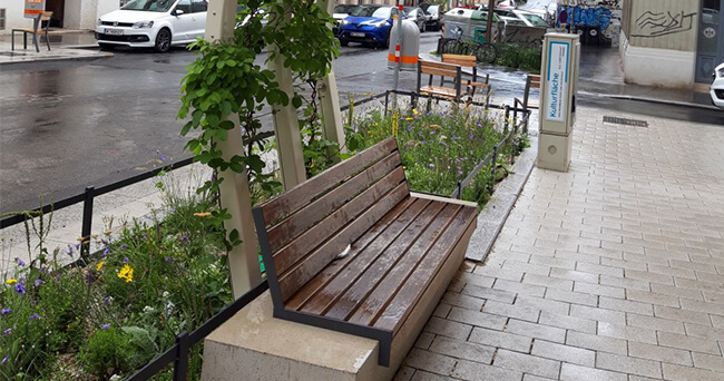 Sitzbank mit Holzlatten an einem Gehweg, eingerahmt von bepflanzten Beeten mit Wildblumen und jungen Bäumen in einer urbanen Straße bei nassem Wetter.