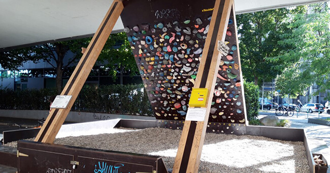 Eine überdachte Kletterwand auf einem öffentlichen Platz, die mit farbigen Klettergriffen ausgestattet ist. Im Vordergrund liegt heller Kies als Fallschutz. Die Umgebung zeigt Bäume und Straßen, die den Platz einrahmen.