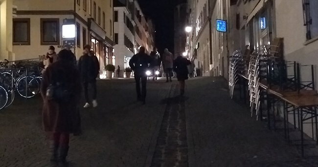 Mehrere Personen gehen am Abend durch eine belebte Altstadtstraße mit Gastronomie und Fahrradständern. Die Szene zeigt eine durchgängige Nutzung des öffentlichen Raums zu später Stunde.