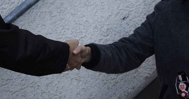 Zwei Personen reichen sich im öffentlichen Raum die Hand. Die linke Person trägt eine dunkle Jacke, die rechte eine graue Jacke mit einer Flasche in der Jackentasche. Der Hintergrund ist eine helle Wand.