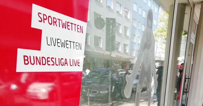 Großes Schaufenster eines Wettbüros mit rotem Werbeschild für Sportwetten, Livewetten und Bundesliga. Im Fenster spiegelt sich eine belebte Straße mit parkenden Autos und Fußgängern.