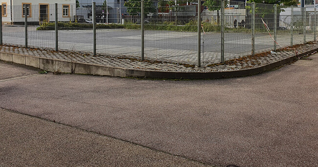 Ein leerstehendes, mit Zaun gesichertes Grundstück in einer Stadt, das möglicherweise für Wohnbebauung vorgesehen ist. Der asphaltierte Vordergrund und die Umgebung lassen auf ein innerstädtisches Areal schließen.