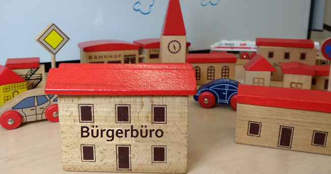 Mehrere Holzspielhäuser mit rotem Dach stehen auf einem Tisch. Im Vordergrund ein Modellhaus mit der Aufschrift „Bürgerbüro“. Im Hintergrund sind ein Bahnhof, eine Kirche und weitere Gebäude aus Holz sichtbar.