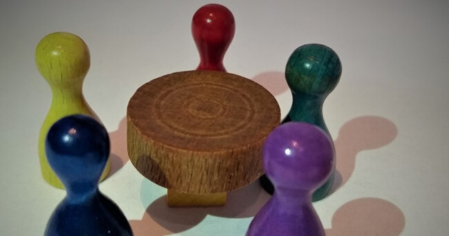 Farbige Spielfiguren aus Holz stehen um einen kleinen runden Holztisch.