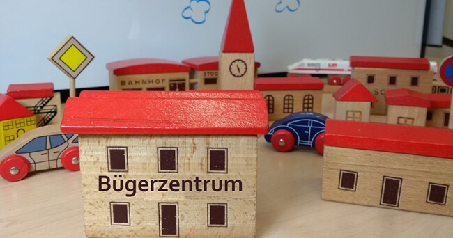 Holzmodell eines Hauses mit der Aufschrift „Bürgerzentrum“ inmitten einer Modellstadt mit weiteren Gebäuden und Autos auf einem Tisch.