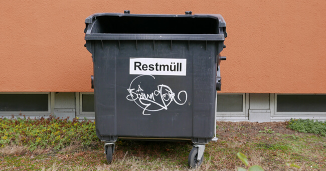 Großes, schwarzes Müllcontainergefäß mit der Aufschrift „Restmüll“ und Graffiti, vor einer orangefarbenen Hauswand platziert.