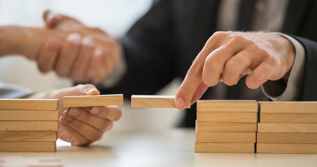 Zwei Personen reichen sich im Hintergrund die Hand, während im Vordergrund Holzbausteine als Brücke zwischen zwei Stapeln gelegt werden.