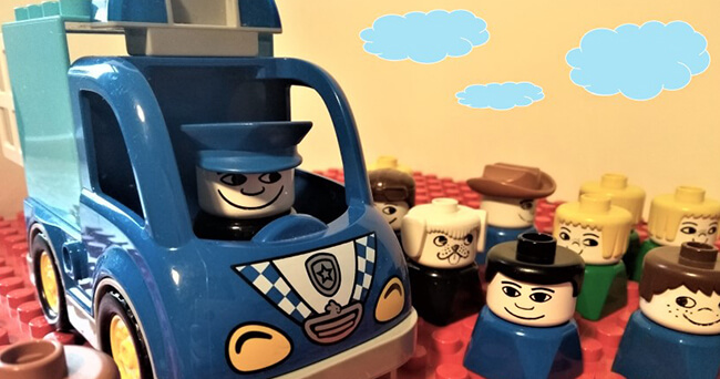 Nahaufnahme eines blauen Spielzeugautos mit einer Polizistenfigur, im Hintergrund mehrere bunte Spielfiguren, auf roter Bauplatte stehend.