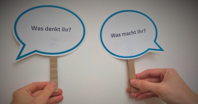 Zwei gebastelte Sprechblasen mit den Fragen „Was denkt ihr?“ und „Was macht ihr?“ in den Händen gehalten.