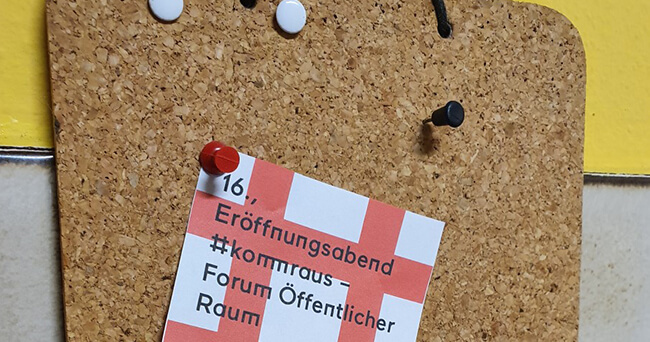 Ein roter Zettel mit der Aufschrift „Eröffnungsabend #kommraus – Forum Öffentlicher Raum“ ist mit einer roten Pinnnadel an einer Korkpinnwand befestigt. Darüber befinden sich weitere Pins.
