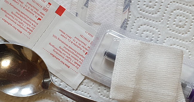 Auf einem weißen Untergrund liegen ein Löffel, sterile Tupfer, eine Spritze und Verbandmaterial.