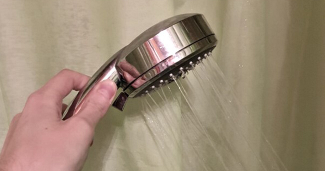 Nahaufnahme einer Hand, die einen Duschkopf hält, aus dem Wasserstrahlen fließen, vor einem hellen Duschvorhang.