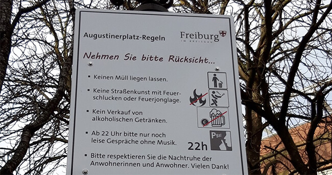 Nahaufnahme eines Hinweisschilds mit Verhaltensregeln am Augustinerplatz in Freiburg, das verschiedene Ge- und Verbote für den öffentlichen Raum auflistet, darunter Müllentsorgung, Straßenkunst, Alkoholverkauf und Lärmschutz.
