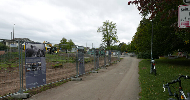 Blick auf eine eingezäunte Baustelle entlang eines Gehwegs, im Hintergrund Baumaschinen, daneben eine offene Grünfläche mit Bäumen.