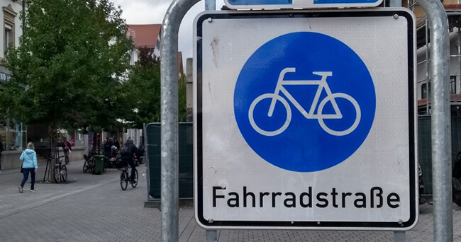 Verkehrsschild „Fahrradstraße“ in einer Innenstadt mit Radfahrenden und Fußgängerinnen und Fußgängern auf einer gemeinsamen Straße bei Tageslicht.