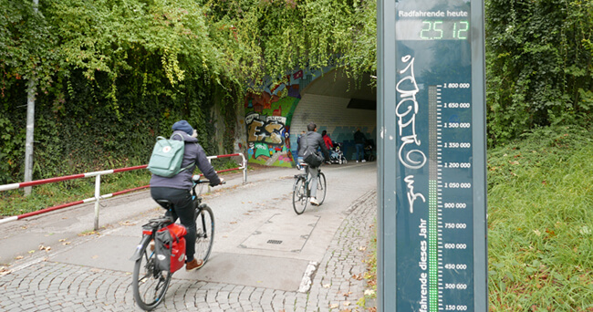 Zwei Radfahrende fahren durch eine mit Graffiti verzierte Unterführung, rechts im Bild steht eine digitale Fahrradzählanlage.