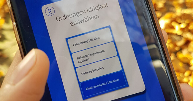 Eine Hand hält ein Smartphone mit geöffneter App zur Auswahl von Ordnungswidrigkeiten wie „Fahrradweg blockiert“ oder „Gehweg blockiert“. Im Hintergrund ist herbstliches Laub zu sehen.