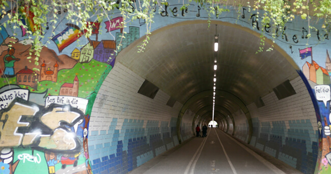 Blick in einen farbig gestalteten, beleuchteten Tunnel mit Wandmalereien und Radweg.