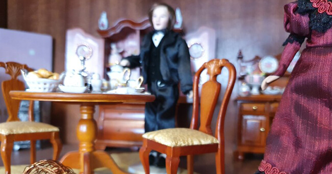 Miniaturdarstellung einer Wohn- und Veranstaltungsszene mit gedecktem Tisch, Stühlen und im Hintergrund stehender Figur in schwarzer Kleidung