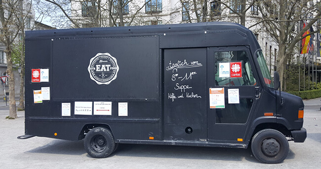 Ein schwarzer Food-Truck mit seitlicher Beschriftung und mehreren Hinweisschildern steht auf einem öffentlichen Platz.