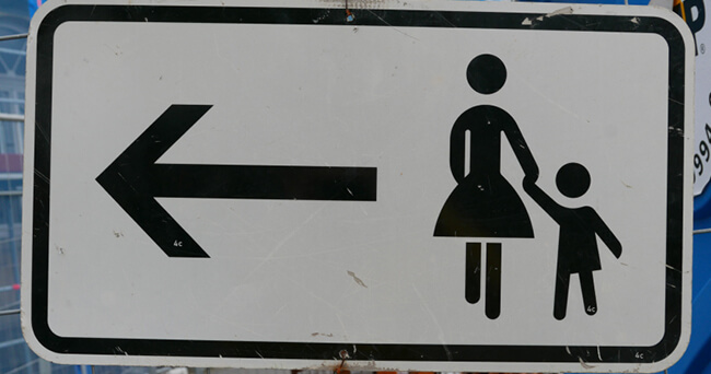 Weiße Hinweistafel mit schwarzem Symbol einer erwachsenen Person, die ein Kind an der Hand hält, und einem schwarzen Pfeil nach links, vor unscharfem Hintergrund.